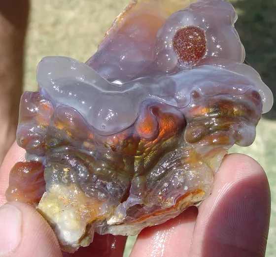 A hand holding rough fire agate: bubbly translucent gray chalcedony over orange, green and gold iridescent layers
