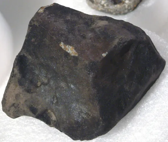 A dark brown-black meteorite fragment with a thin dark crust and a small light-colored chip